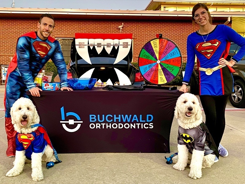 Doctor Buchwald two dogs and team member dressed as superheroes next to table with prize wheel